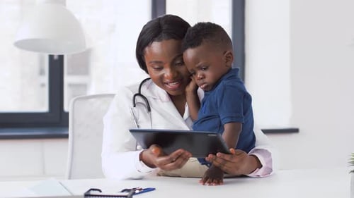 Happy african american pediatrician shows tablet to baby boy patient at clinic