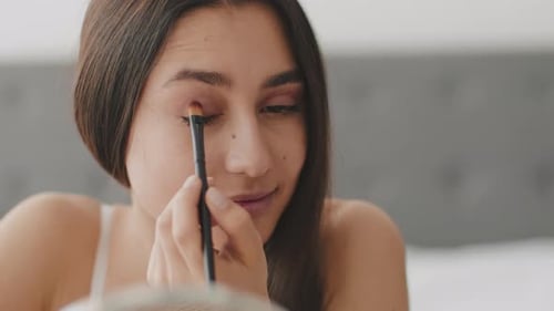 Woman applies eyeshadow makeup in bedroom, close up