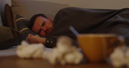 a man lying on a couch blows his nose and is sick. In the foreground tea and handkerchiefs