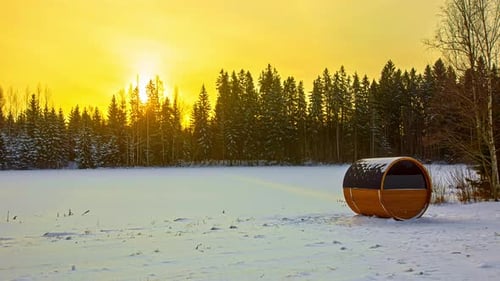 Barrel Sauna On Snowy Field With Coniferous Forest In Background And Bright Yellow Sunset Sky. - tim