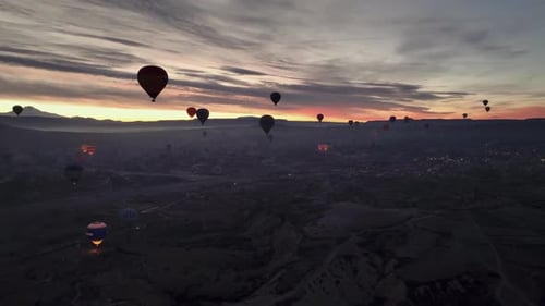 Drone view of numerous hot air balloons flying low over the ground at sunrise in Cappadocia, with mo