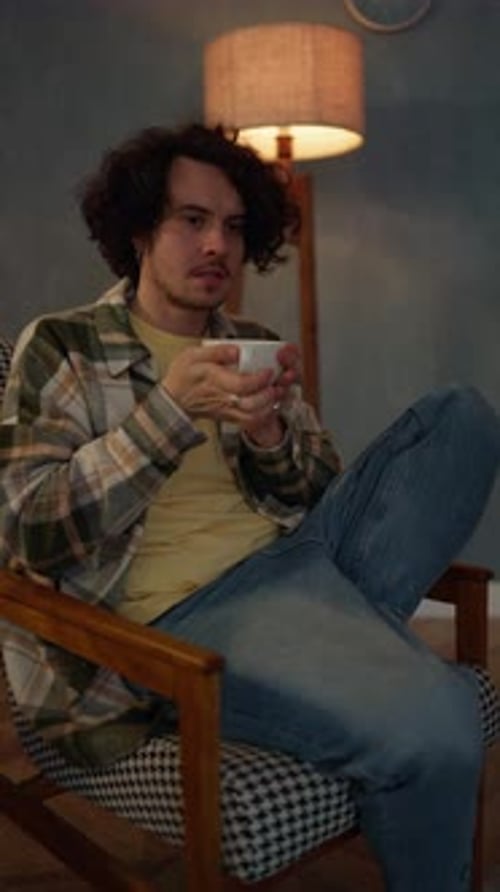 Vertical Video of a Brunette Guy with Curly Hair Brings a Cup of Tea to His Face with Both Hands and