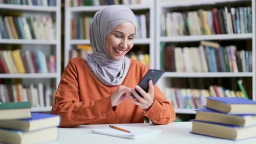 Muslim female student in hijab using smartphone in campus library space. Happy girl looking