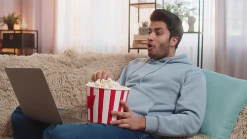 человек смотрит фильм на диване с ноутбуком и попкорном