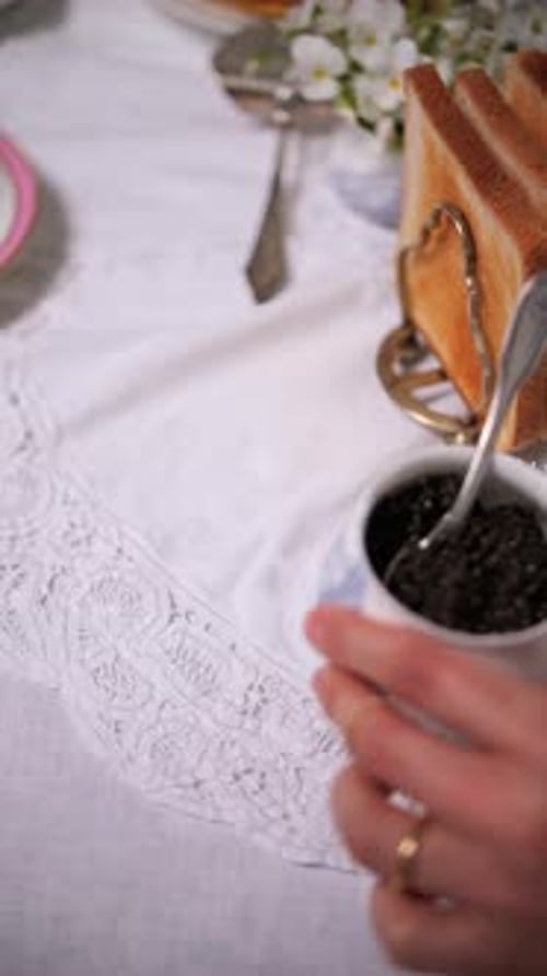 Woman's Hand Placing Black Caviar on Table with Toast and Floral Decor