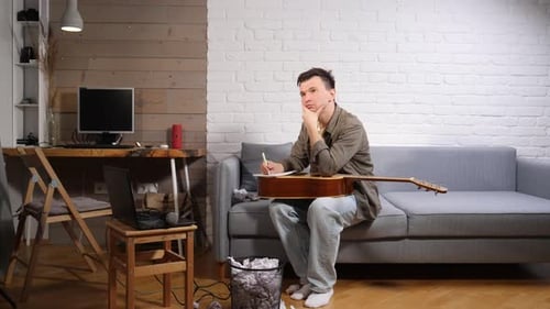 Young Man Sitting on a Sofa with an Acoustic Guitar Covering His Face with Hands Surrounded By