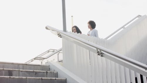 Diverse female friends pausing to gaze skyline from overpass, holding coffee, descending stairs