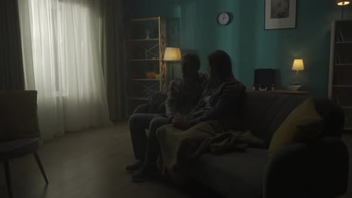 A Couple is Sitting on a Sofa in the Dark in the Room A Man Listens to a Woman Holds Her Hand Hugs