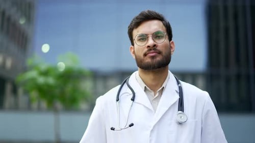 Portrait of serious doctor in a white coat looking at camera, standing outside near hospital build