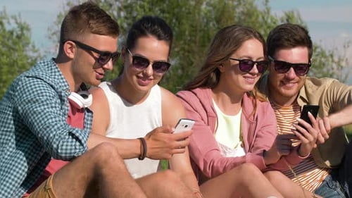 Smiling friends with smartphones enjoying summer vacation in the park