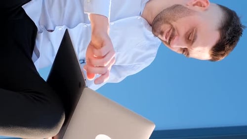 A Handsome Man is Sitting Outside Near an Office Center and Working on a Laptop Vertical Video