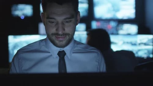 Focused security officer is working on a computer in a dark monitoring room filled with display scr