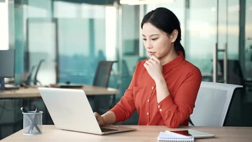Serious asian female employee is using laptop sitting at workplace in business office. Busy woman