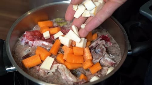 Person adding chopped parsnips to bone broth beginning to boil in pot.
