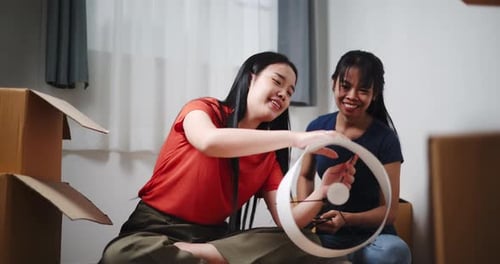 Two Asian siblings sit on the floor and help assemble a floor lamp in their new home.