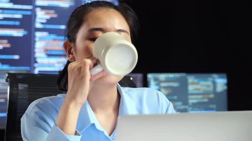 Une programmeuse asiatique boit du café tout en écrivant du code sur un ordinateur portable à l'aide de plusieurs moniteurs