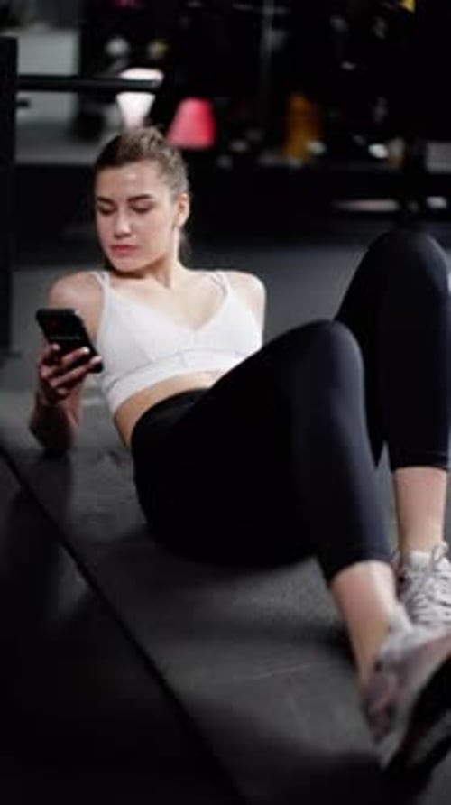 Relaxed Athletic Woman in Workout Gear Using Smartphone on Gym Floor Taking a Break From Fitness