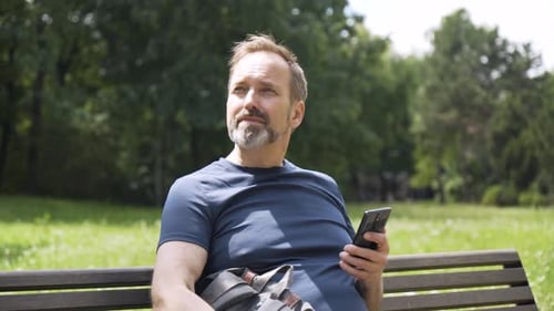 Un apuesto hombre caucásico de mediana edad trabaja en un teléfono inteligente con una sonrisa mientras se sienta en un banco