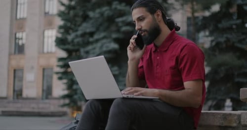 Joyful Arab Man Freelancer Working Outdoors Using Laptop Typing and Talking on Mobile Phone in City
