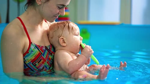 Woman is holding a little cute baby boy in the swimming pool. Lovely kid is chewing the toy.
