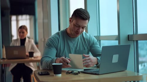 Busy young man sitting at desk looking through the paper notebook. Businessman working on laptop.