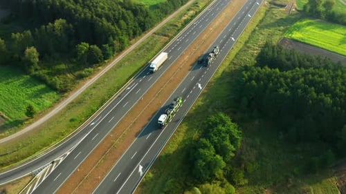 Trucks Loaded with Cars Move Along Rural Road Passing Green Fields