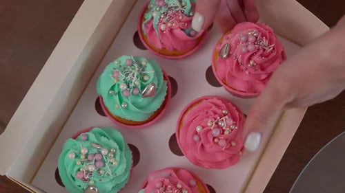 Top View Closeup Box with Baked Decorated Muffins and Female Caucasian Hand Placing Cupcake Inside