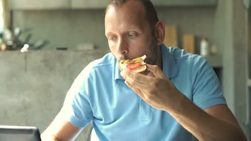 Man Eating Sandwich While Using Laptop Indoors