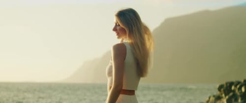 Woman poses elegantly by the ocean with sunlight illuminating her profile against the cliffs.