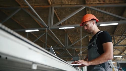 Worker in uniform stands in the factory and typing on laptop