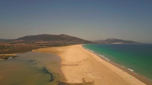 AERIAL ZOOMING OUT. Spit of sand along European coastline with mountain range in background.