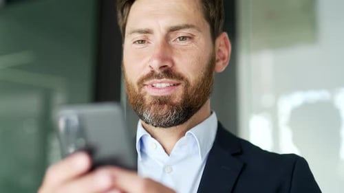 Smiling businessman in formal suit is using smartphone at workplace in business office.