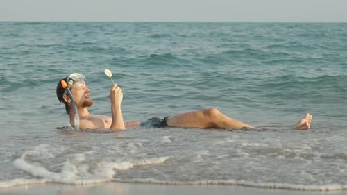 Un homme masqué et tuba est allongé au bord de la mer et lèche une sucette colorée sur un bâton. Il était
