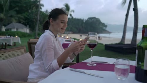 Woman Texting On Smartphone Sitting In Beach Restaurant In Evening