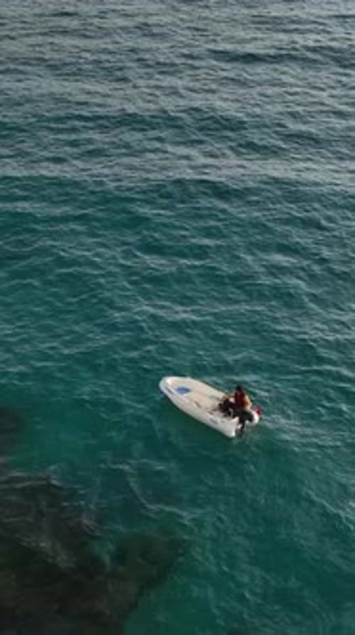 Vertical Video Lonely Fisherman in an Inflatable Boat Floating on a Clear Blue Sea Under Bright