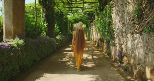 Walking through a shaded garden path, the woman in a yellow dress and straw hat enjoys the greenery