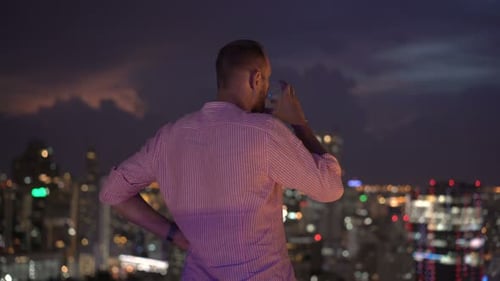 Young man enjoys a cocktail and city view on a rooftop terrace at night