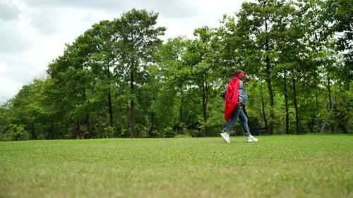 Happy girl are playing superhero. Kid run across green field in red cloak
