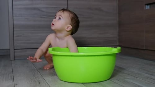 Naked little toddler sits behind the green tub indoors.