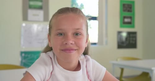 Preteen girl smiling then speaking to camera at desk in class, giving presentation in pink pinafore