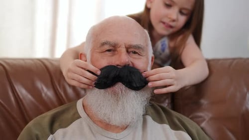Front View Closeup of Positive Senior Bearded Man with Blurred Girl Placing Toy Black Mustache on