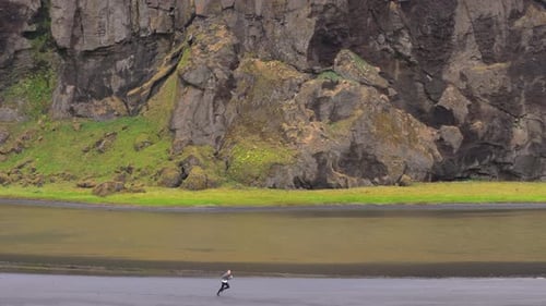 Drone Capturing Man Running on Black Shore at the Foot of Mossy Rock Iceland