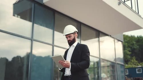 Serious Bearded Businessman in Suit and Safety Helmet Walks Down the Building Area Actively Using