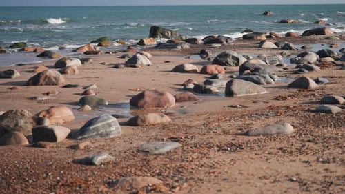 Pedras e seixos cobrem a praia arenosa enquanto as ondas rolam lentamente ao fundo.