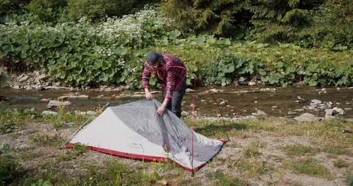 A Review of a Guy in a Red Plaid Shirt Who Assembles a Tent on His Hike Near a Mountain River in the