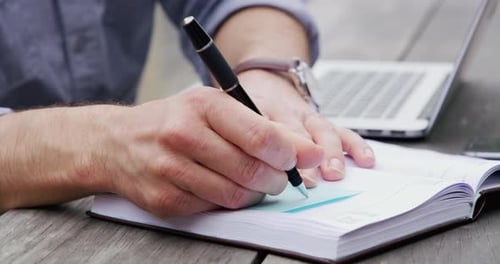 Man Writing in Notebook with Laptop on Desk