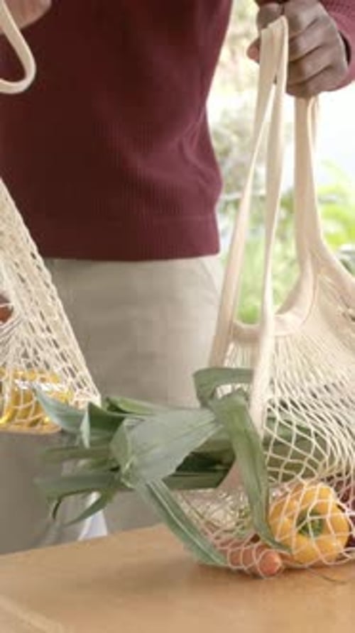 Vídeo vertical: Llega a casa, un hombre con un jersey rojo coloca bolsas de red sobre una mesa de madera con verduras y tarros