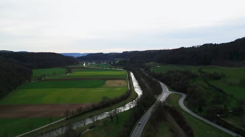 Drone flying over green agriculture fields, river and speed highway surrounded by hills.