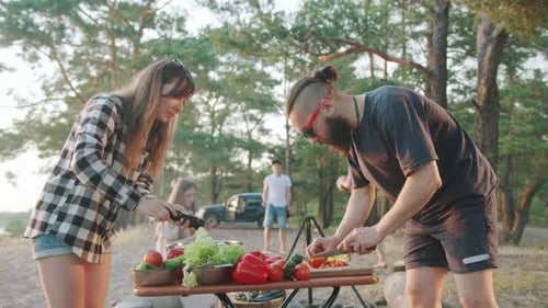 Hipster Man and a Young Beautiful Girl Prepare a Picnic Lunch While Friends Play Ball Near a Pine