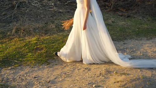 Slow-Motion Close-Up of a Bride in a White Wedding Dress Walking in Sunset Light Holding Dry Grass